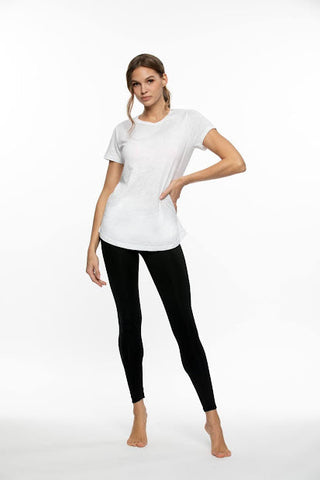 A woman wearing a white t-shirt and black leggings is barefoot and has one hand on her hip, while looking at the camera.