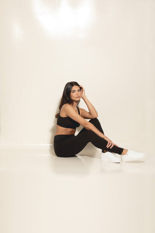 A person wearing a black Felina sports bra, leggings, and white shoes, sitting in front of a beige backdrop.
