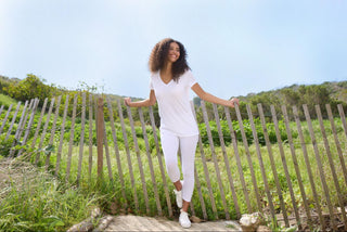 A smiling woman with curly hair, wearing a white V-neck t-shirt, white leggings, and white sneakers, leans against a rustic wooden fence in a sunny, grassy field.
