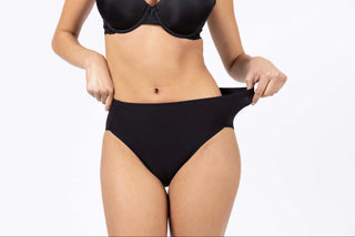 A close-up shot from the chest down of a person wearing a black underwire bra and high-waisted black underwear, pulling on the band of the right side of the image, against a plain white background.
