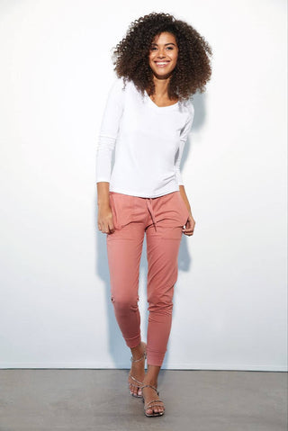 A person smiling at the camera and wearing Felina’s pink Velvety Soft Joggers, a white, long-sleeve top, and sandals, standing in front of a white backdrop.